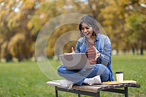 Excited Young Arab Female Student Using Laptop While Sitting On Bench Outdoors