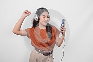 Excited young Asian woman in brown shirt raising clenched fist gesture while listening to music from smartphone using wireless