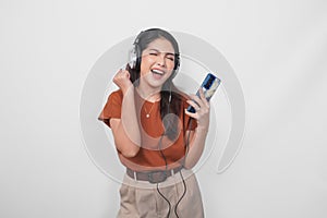 Excited young Asian woman in brown shirt raising clenched fist gesture while listening to music from smartphone using wireless