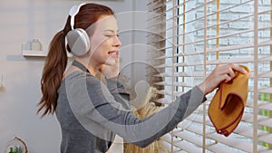 Excited young asian woman with headphones singing and dancing while dusting a window in living room.