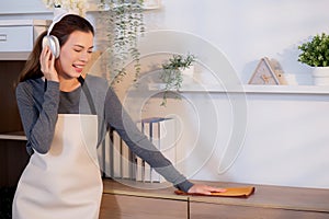 Excited young asian woman with headphones singing and dancing while dusting a wooden cabinet in living room.