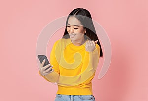 Excited young Asian woman received a good message on the mobile phone, happy with great news standing on pink background