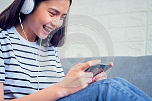 Excited young Asian woman sitting on sofa wear white headphone on the head and playing games.