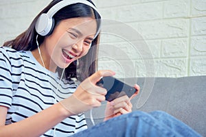 Excited young Asian woman sitting on sofa wear white headphone on the head and playing games.