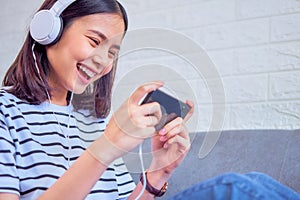 Excited young Asian woman sitting on sofa wear white headphone on the head and playing games on smartphone in the living room.