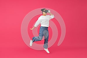 Excited young asian woman wearing VR headset jumping on pink background