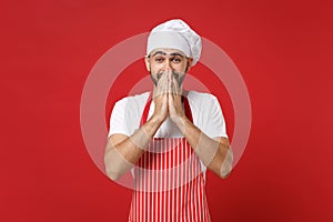 Excited young bearded male chef cook or baker man in striped apron white t-shirt toque chefs hat posing isolated on red