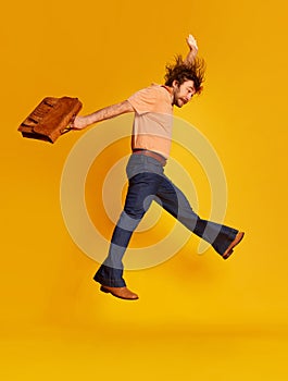 Excited young bearded man, hippie in stylish sunglasses and flared jeans running jumping over yellow background. Retro