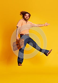 Excited young bearded man, hippie in stylish sunglasses and flared jeans running jumping over yellow background. Retro