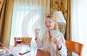 An excited young blonde of European origin, dressed in a white blouse, holds dollar bills and clenches her fist