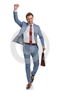 Excited young businessman with arm up holding suitcase and celebrating