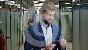 Excited young businessman rejoices at message on his smartphone, standing at office. Happy male looking at mobile phone