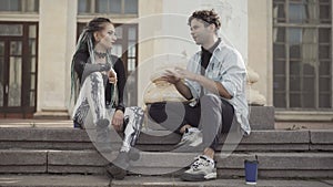 Excited young Caucasian hippie sitting on urban stairs with goth woman and talking. Portrait of relaxed millennial