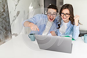 Excited young couple celebrating success, reading good news in email, looking at laptop screen, standing in modern kitchen at home