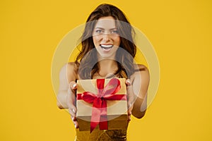 Excited young european woman in festive dress giving box with present, stretching gift to camera on yellow background