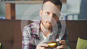 A excited young gamer is sitting on a couch and playing video games on a console. The man is playing with a wireless