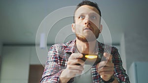 A excited young gamer is sitting on a couch and playing video games on a console. The man is playing with a wireless