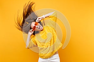 Excited young girl in fur sweater holding heart orange glasses fooling around in studio jumping with fluttering hair