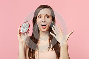 Excited young girl in light clothes holding in hand half of fresh ripe pitahaya dragon fruit isolated on pink pastel