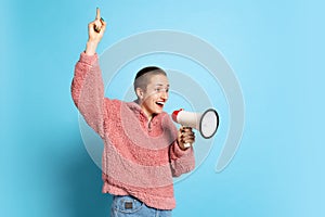 Excited young girl, student with short hair wearing pink hoodie shouting at megaphone isolated on blue background