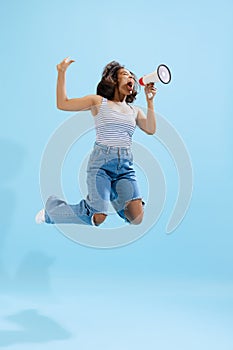 Excited young girl, student wearing casual style clothes jumping and shouting at megaphone isolated on light blue