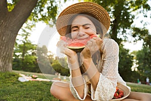 Excited young girl in summer hat having a picnic at the park, sitting on a grass,