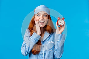 Excited young girl wake up on time, holding red cute clock set alarm and smiling amused, wearing nightwear, pyjama and