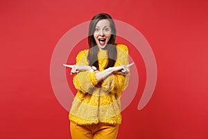 Excited young girl in yellow fur sweater with opened mouth holding hands crossed pointing index fingers aside isolated