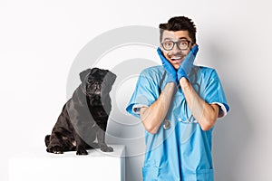 Excited young male doctor veterinarian admiring cute pet sitting on table. Cute black pug dog waiting for examination at