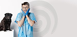 Excited young male doctor veterinarian admiring cute pet sitting on table. Cute black pug dog waiting for examination at