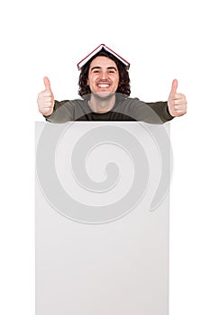 Excited young man, with a book on his head, shows thumbs up gesture, standing behind a blank announcement banner. Joyful guy looks