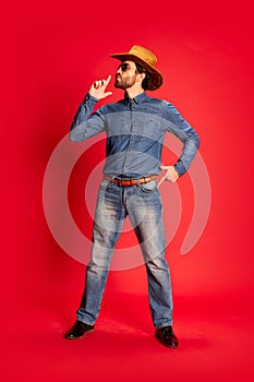 Excited young man in cowboy style outfit having fun, posing isolated over red background. Model in cowboy hat and denim