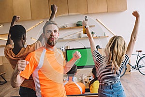 excited young man doing yes gestures and looking at camera while his friends celebrating and watching soccer match on tv screen