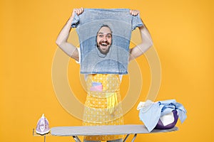 Excited young man househusband in apron ironing clean clothes on board while doing housework isolated on yellow wall