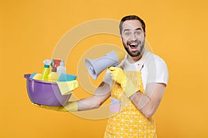 Excited young man househusband in apron rubber gloves hold basin with detergent bottles washing cleansers doing