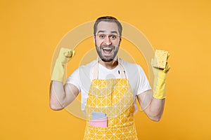 Excited young man househusband in apron rubber gloves hold cleaning sponge rag while doing housework isolated on yellow