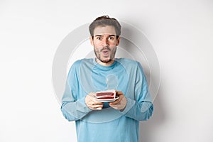 Excited young man making wish on birthday cake with lit candle, celebrating bday, standing on white background