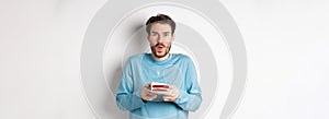 Excited young man making wish on birthday cake with lit candle, celebrating bday, standing on white background