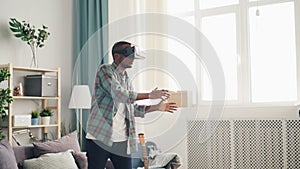 Excited young man is playing game wearing virtual reality glasses at home moving arms and body standing in light room