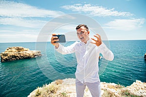 Excited young man is taking photo of himself on mobile phone. He is standing on cliff near the sea and scream