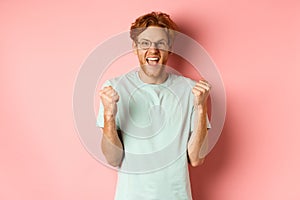 Excited young man winning prize, shouting with joy and triumph, making fist pump and saying yes, standing over pink