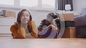 Excited young multiracial couple lying on the floor of their new appartment. Cardboard boxes around and woman holding