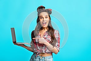 Excited young pinup womanin retro outfit holding laptop and pointing at screen, looking at camera over blue background