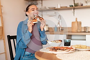 Excited young pregnant lady enjoying pizza, biting two tasty slices, sitting at table in kitchen interior, copy space