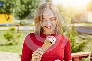 Excited young pretty female with straight light hair, having freckled face looking at ice-cream with great happiness standing in p