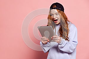Excited young redhead girl, student in hoodie and cap isolated on pink studio background. Fashion, human emotions