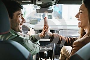 Excited young spouses celebrating purchase of new car, sitting inside modern auto, high fiving to each other at