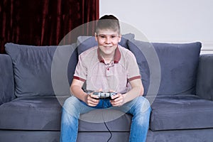 Excited young teenager boy gamer playing video games with joystick, sitting on couch at home