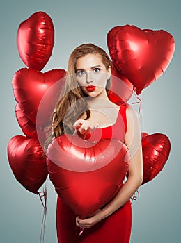 Excited young woman blowing kiss and holding balloons red heart. Beautiful girl with red lips makeup, long curly hair