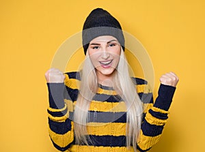 Excited young woman with clenched fists, model wearing woolen cap and sweater, isolated on yellow background. Yes concept. Good